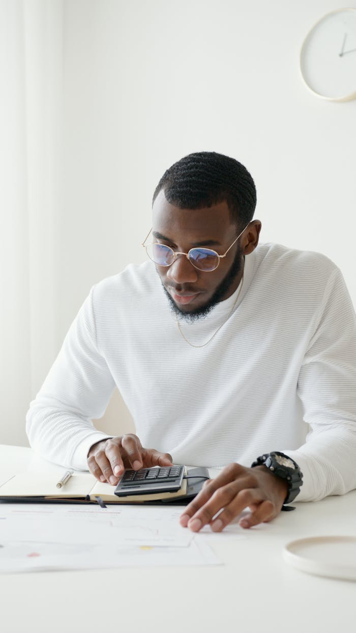 Man in glasses working on calculations at desk in bright office setting.